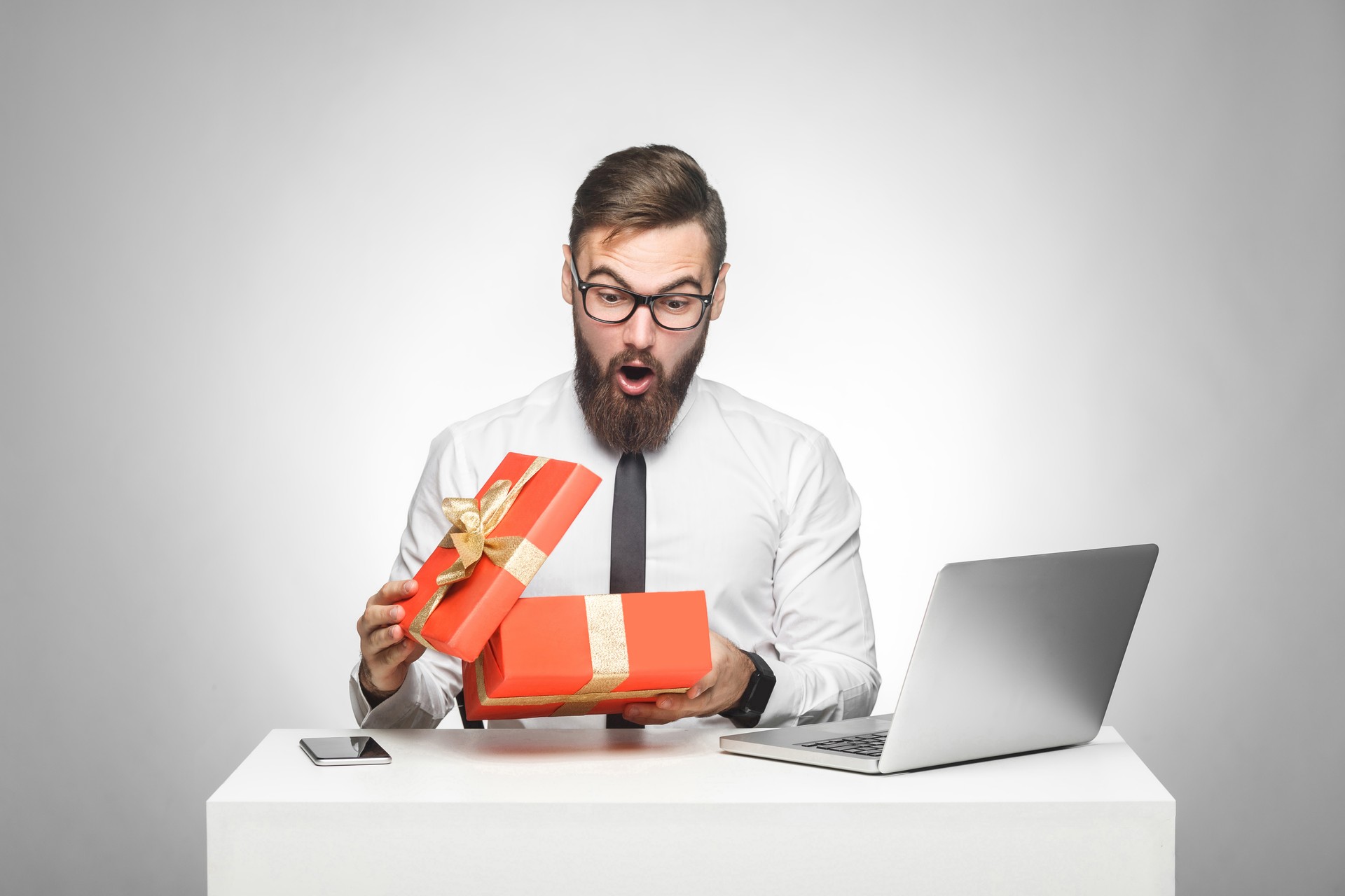 Wow! Suprised young manager in white shirt and black tie are sitting in office and unboxing present with shocked face, big eyes and opened mouth.