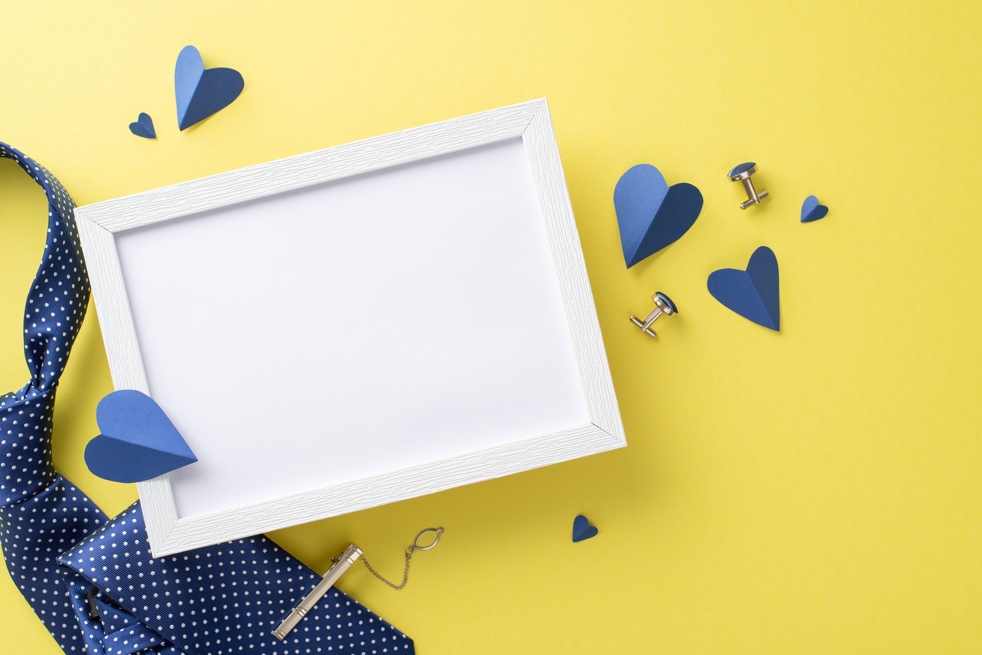 Celebrate Father's Day with this top-view composition. Hearts, accessories, photo frame, a blue necktie, and cufflinks, on a yellow backdrop, featuring an empty frame for text or picture